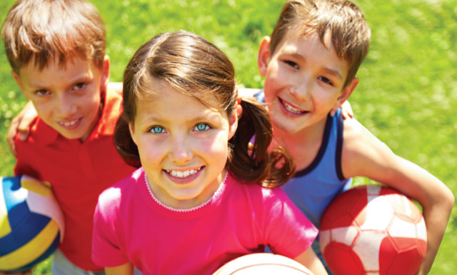 Three children smiling up at the camera standing in a field.