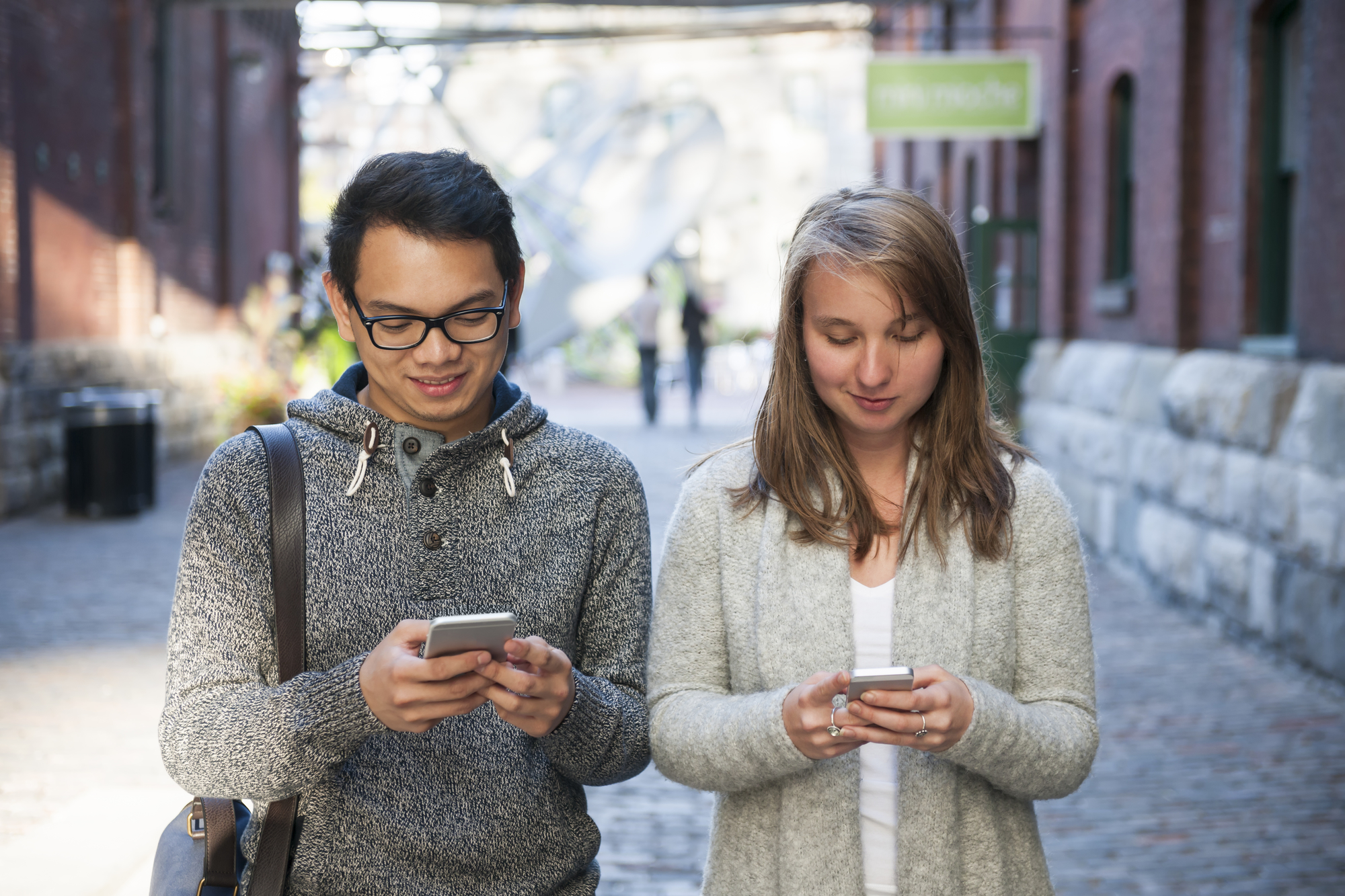 Two young people looking into smartphones while walking on city street.