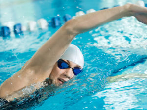 Young woman in goggles and cap swimming front crawl stroke style in the blue water indoor race pool.