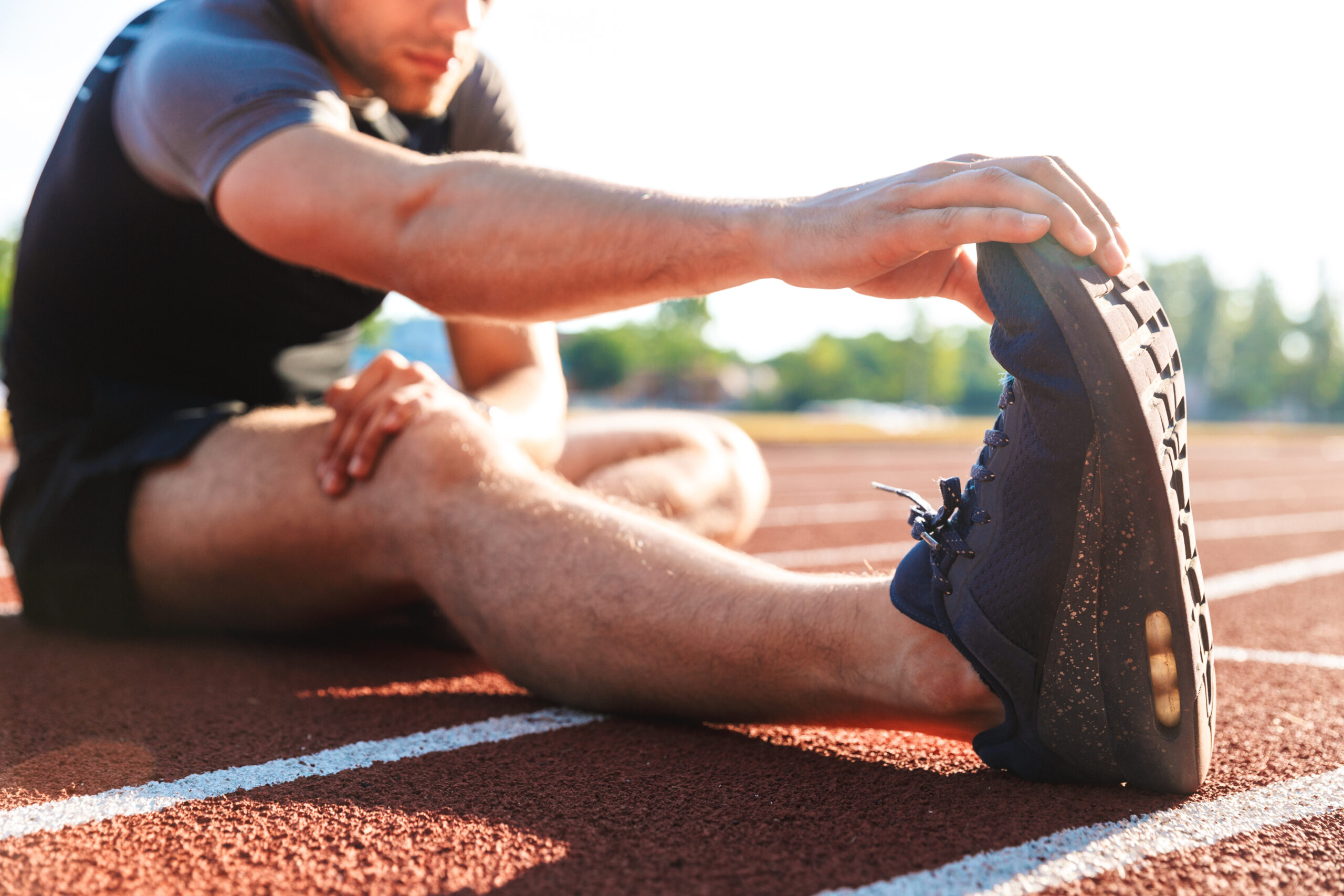 close up of male athlete sitting on track stretching, holding his foot with his hand